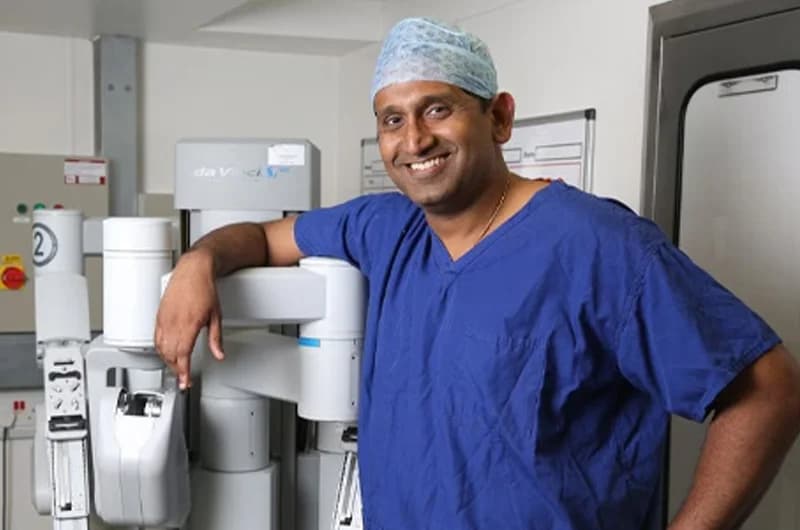 Professor Prasanna Sooriakumaran in scrubs beside a robotic surgical system